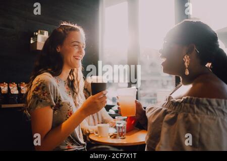 Seitenansicht der fröhlichen multirassischen jungen Casual Frauen lachen und Kaffee trinken, während man am Fenster im Cafe bei Sonnenuntergang sitzt Stockfoto