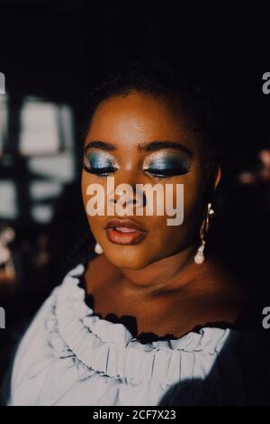 Portrait von hintergrundbeleuchteten schönen kurvigen schwarzen jungen Frau mit hellen Make-up in off-shoulder Kleid Blick nach unten Stockfoto