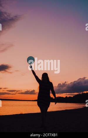 Rückansicht einer anonymen Frau mit Ballon und einem Spaziergang entlang der Küste bei schönem Sonnenuntergang Stockfoto