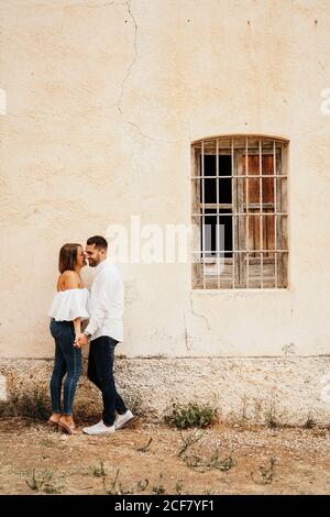 Vollkörper glücklicher Mann und Frau, die Hände und berührende Nasen halten, während sie in der Nähe einer alten Hütte mit geriebenem Fenster stehen Stockfoto