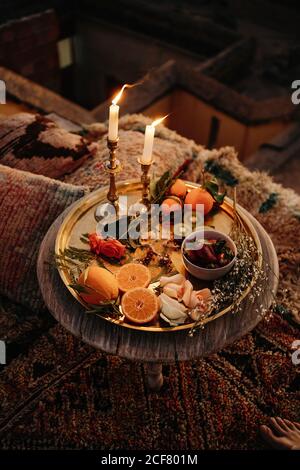 Von oben Zusammensetzung von verschiedenen frischen Früchten und Pflanzen serviert Auf goldenem Tablett mit brennenden Kerzen auf runder Holzplatte Im marokkanischen Stil Stockfoto