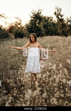 Friedliche junge Frau in weißem romantischen Kleid steht und berührt hohes Gras mit Feldblumen und schaut auf die Kamera Stockfoto