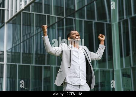 Schöner afroamerikanischer Geschäftsmann feiert den Sieg und hebt die Hände auf den Hintergrund Glaswand Stockfoto