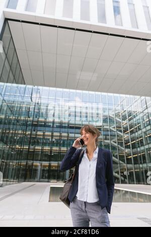 Glückliche Geschäftsfrau, die die Hand in der Tasche hält und das Smartphone benutzt, während sie vor dem Business Center auf der Straße der Stadt steht Stockfoto