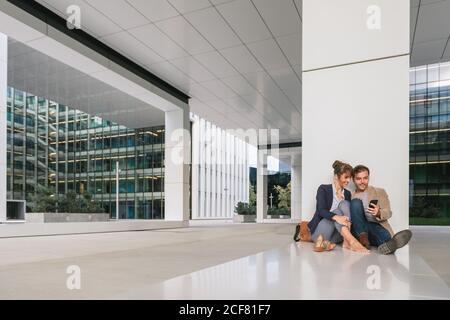 Fröhliche Paar umarmen einander beim Surfen auf einem Smartphone Sitzen Sie vor einem modernen Gebäude an einer Stadtstraße zusammen Stockfoto