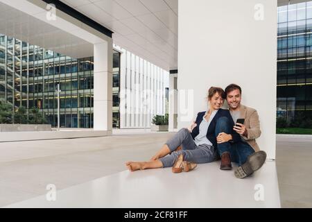 Fröhliche Paar umarmen einander beim Surfen auf einem Smartphone Sitzen Sie vor einem modernen Gebäude an einer Stadtstraße zusammen Stockfoto