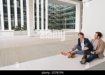 Fröhliche Paar umarmen einander beim Surfen auf einem Smartphone Sitzen Sie vor einem modernen Gebäude an einer Stadtstraße zusammen Stockfoto