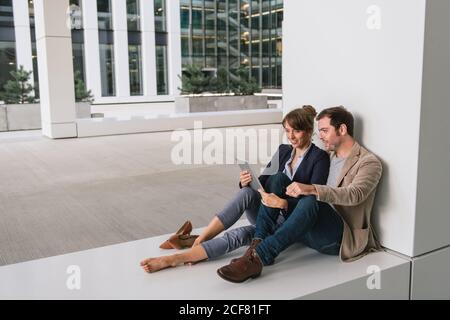 Fröhliche Paar umarmen einander beim Surfen auf einem Tablet Sitzen Sie vor einem modernen Gebäude an einer Stadtstraße zusammen Stockfoto