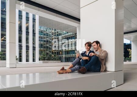 Fröhliche Paar umarmen einander beim Surfen auf einem Tablet Sitzen Sie vor einem modernen Gebäude an einer Stadtstraße zusammen Stockfoto