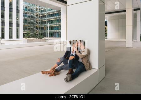 Männlich Manager umarmt und küssen Freundin während sitzen außerhalb Büro Gebäude an der Stadtstraße nach der Arbeit Stockfoto