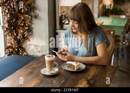 Blonde junge fröhliche Frau in Freizeitkleidung Eingabe Nachricht auf Smartphone beim Sitzen am Tisch mit Kaffee und Dessert in Gemütliches Café Stockfoto