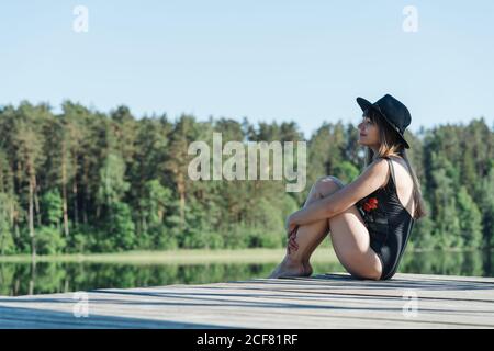 Seitenansicht einer glücklichen jungen Frau in schwarzem Badeanzug und Hut, die auf einem hölzernen Pier sitzt und den Blick auf den See am klaren blauen Himmel und auf dem Waldhintergrund bewundert Stockfoto
