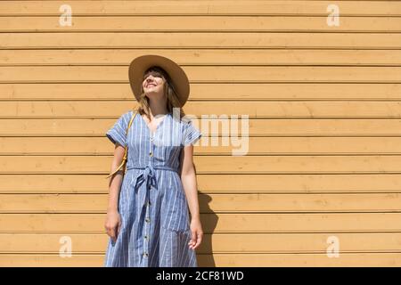 Eine hübsche lächelnde Frau mit Strohhut und Sundress, die mit geschlossenen Augen vor einer Holzwand steht Stockfoto