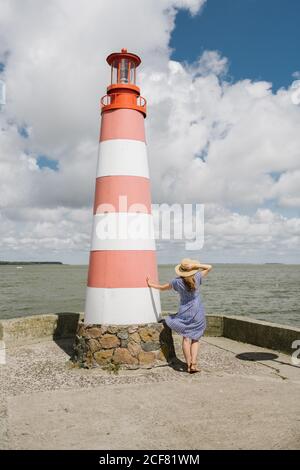 Rückansicht einer Frau in Strohhut und wehendem Kleid, die an windigen Tagen in der Nähe des gestreiften Leuchtturms am Ufer steht Stockfoto