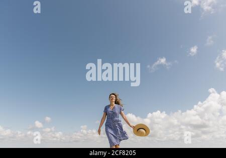 Inhalt Erwachsene Frau mit wehenden Haaren und in Sundress zu Fuß mit Strohhut in der Hand auf bewölktem Himmel Hintergrund Stockfoto