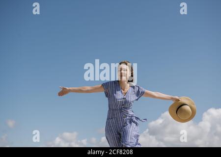 Inhalt Erwachsene Frau mit wehenden Haaren und in Sundress zu Fuß mit Strohhut in der Hand auf bewölktem Himmel Hintergrund Stockfoto