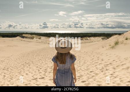 Erwachsene Frau in legerem Kleid und Strohhut, die an heißen Sommertagen auf Sanddüne steht Stockfoto