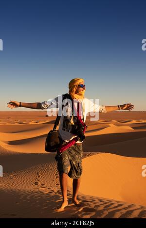 Nicht erkennbarer Tourist mit ausgestreckten Armen, die gegen den hellen, wolkenlosen Sonnenuntergang stehen Himmel in der Wüste Stockfoto