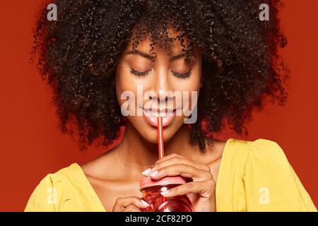 Nahaufnahme einer verträumten jungen afroamerikanischen Dame mit Locken Haare halten roten Glas mit Stroh und genießen Getränk auf Roter Hintergrund Stockfoto