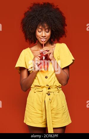 Verträumte junge afroamerikanische Dame mit lockigen Haaren hält rot Glas mit Stroh und Getränk auf rotem Hintergrund genießen Stockfoto