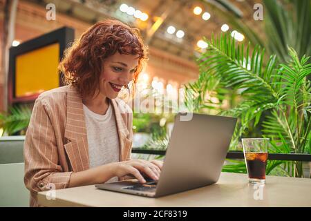 Glückliche junge Geschäftsfrau, die mit einem kalten Getränk im Café am Tisch sitzt und einen Laptop für die Arbeit benutzt Stockfoto