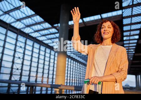 Hübsche lächelnde junge Frau mit dem Koffer, der die Hand für Auf Wiedersehen winkt, während sie am Bahnhof steht Stockfoto