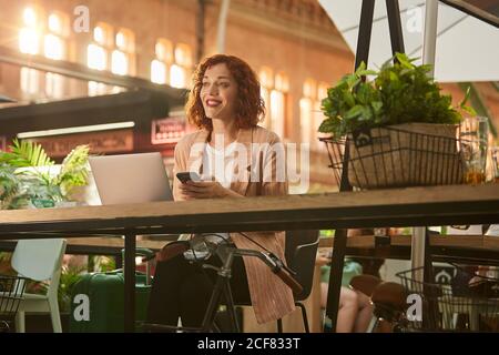 Fröhliche junge Frau, die im Café am Laptop arbeitet und im Telefon surft Stockfoto
