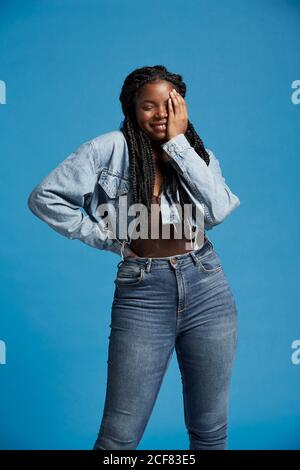 Positive schöne afroamerikanische Frau mit Zöpfen im Denim-Outfit lächelnd mit geschlossenen Augen vor blauem Hintergrund Stockfoto