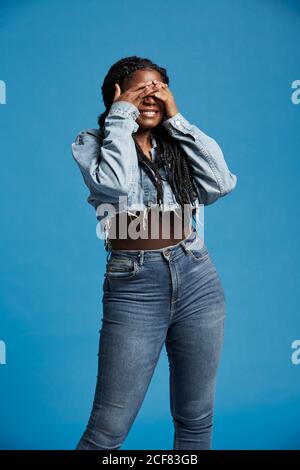 Positive schöne afroamerikanische Frau mit Zöpfen im Denim-Outfit lächelnd und bedeckendes Gesicht mit Händen vor blauem Hintergrund Stockfoto