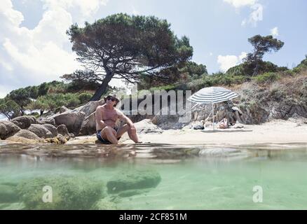 Entspannender älterer Mann im Hut, der am Sandstrand mit frischem Wasser liegt, das in hellem Sonnenlicht ruht, Chalkidiki, Griechenland Stockfoto
