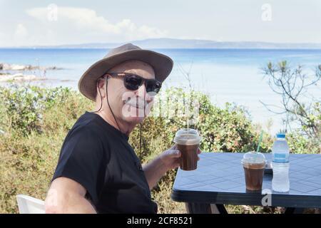 Lächelnder alter Mann mit Sonnenbrille und Hut, hält ein Plastikglas und schaut auf die Kamera, die am Tisch am Meer in Chalkidiki, Griechenland, sitzt Stockfoto