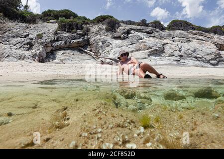 Entspannender älterer Mann im Hut, der am Sandstrand mit frischem Wasser liegt, das in hellem Sonnenlicht ruht, Chalkidiki, Griechenland Stockfoto
