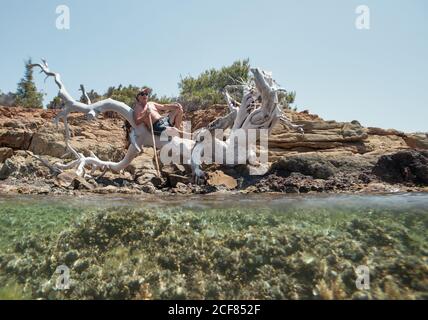 Gebräunter älterer Mann in Sonnenbrille und Hut, der Ruhe auf altem trockenen weißen Baum nimmt, der über klarem Wasser der Bucht, Chalkidiki, Griechenland hängt Stockfoto