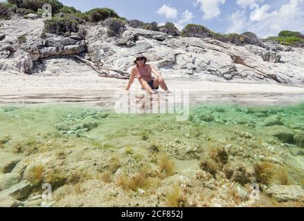 Entspannender älterer Mann im Hut, der am Sandstrand mit frischem Wasser liegt, das in hellem Sonnenlicht ruht, Chalkidiki, Griechenland Stockfoto