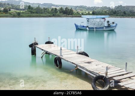 Blaues Boot, das in der Nähe eines kleinen hölzernen Piers mit schwarzen Reifen an den Seiten bei sonnigem Tag segelt, Chalkidiki, Griechenland Stockfoto