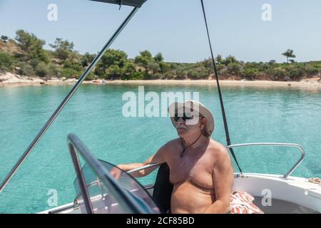Älterer Mann in Hut und Sonnenbrille auf schwarz-weißem Boot in ruhigen türkisfarbenen Wasser von Chalkidiki, Griechenland sitzen Stockfoto