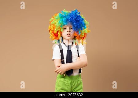 Kleiner Junge in Clown Perücke schmunzelt und Spaß haben. Fröhlicher Clownsjunge mit großer bunter Perücke. Geburtstagskind. Kleiner Clown Junge mit bunten Haaren. Positiv Stockfoto