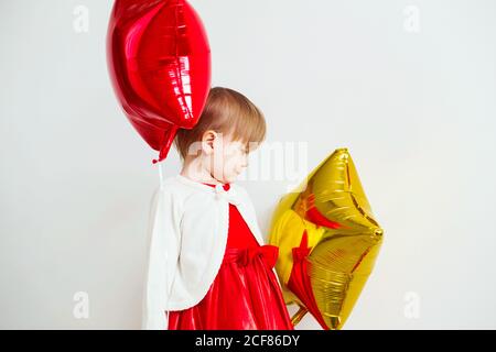 Nettes kleines Mädchen spielt mit sternförmigen Ballons vor weißem Hintergrund. Mädchen schaut und freut sich über ihre Reflexion in Folienballon. kinderlauge Stockfoto