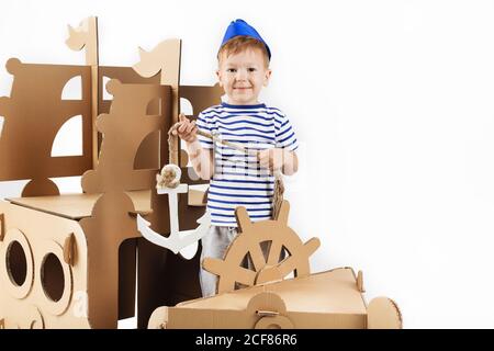 Kleiner Junge spielt mit Pappschiff auf weißem Hintergrund. Frohe Kinder. Nettes Kind als Seemann gekleidet. Kindheit. Fantasie, Fantasie. Stockfoto