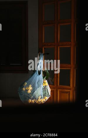 Wiederverwendbarer weißer Saitenbeutel mit frischen Loquatfrüchten auf Ästen Hängen an Knopf der Holztür im Zimmer mit dunkel Leicht Stockfoto