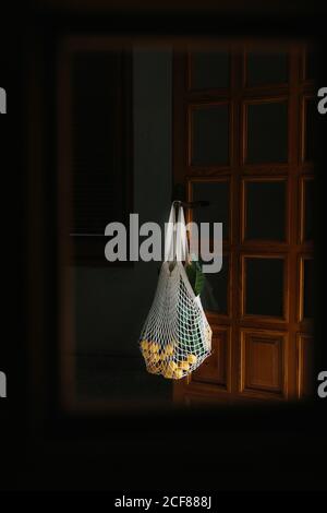 Wiederverwendbarer weißer Saitenbeutel mit frischen Loquatfrüchten auf Ästen Hängen an Knopf der Holztür im Zimmer mit dunkel Leicht Stockfoto