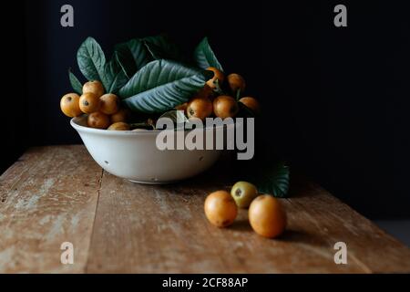 Weiße Keramikplatte mit leckeren natürlichen Loquatfrüchten auf Zweig Mit frischen grünen Blättern auf Holz schäbigen Schreibtisch platziert Stockfoto