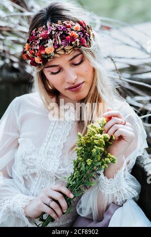 Schöne junge Frau in weißem Kleid und Blumenkranz berühren Bouquet von kleinen gelben Blumen während der Ruhe im Garten am Hochzeitstag Stockfoto
