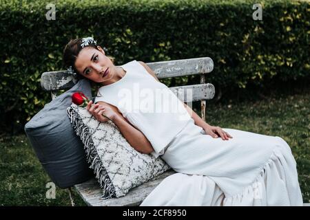 Attraktive junge Dame in weißem Kleid und mit roter Rose Lehnen Sie sich auf weichen Kissen und blicken Sie weg, während Sie sich darauf ausruhen Schäbige Bank am Hochzeitstag im Park Stockfoto