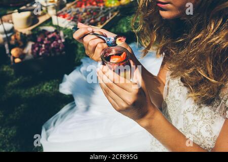 Von oben Blick auf anonyme junge Dame in elegantem Weiß Hochzeitskleid Essen köstliche süße Erdbeere Dessert mit Löffel während Mittagessen im Freien Stockfoto