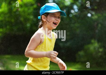 Kleines Mädchen lachend gesprüht, von Wasser im Hinterhof, Garten, Wassertropfen in der Luft spritzt. Sommeraktivitäten, draußen spielen Stockfoto