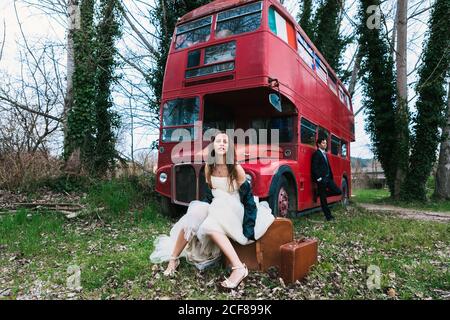 Braut im Hochzeitskleid und Lederjacke sitzt auf Retro Koffer und Bräutigam im Anzug auf rotem Doppeldecker lehnt Bus im Wald verlassen Stockfoto