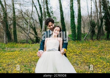 Happy Bräutigam in eleganten Anzug umarmt lächelnde Braut mit gelb Löwenzahn auf blühender Wiese während des Hochzeitstages Stockfoto