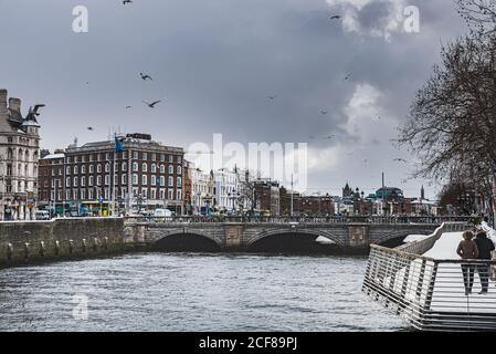 Entspannte Menschen laufen auf der verschneiten Liffey Promenade, während Möwen in der Szene fliegen. Ruhiges Paar genießen die graue Skyline der Innenstadt. Dublin Stockfoto