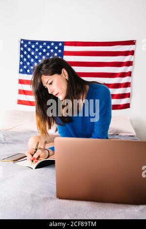 Fokussierte junge Frau in Freizeitkleidung Lesen von Notizen in Tagebuch Während Sie auf dem Bett in der Nähe des Laptops sitzen, während Sie ferngesteuert arbeiten Zu Hause Stockfoto